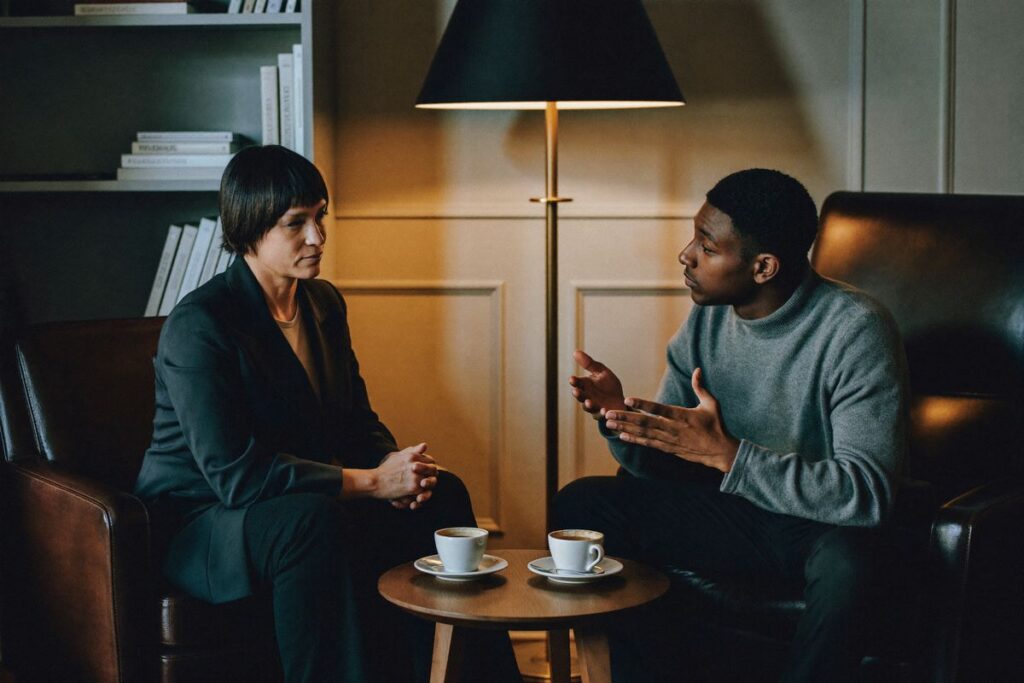 An operations lead listens as a younger colleague explains his work over coffee in an office lounge
