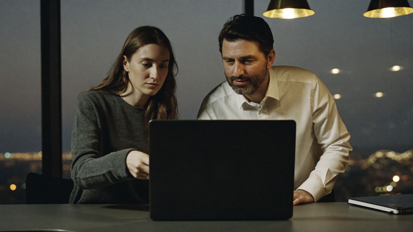 Two colleagues work through a task together on a laptop at a standing desk by a window at dusk