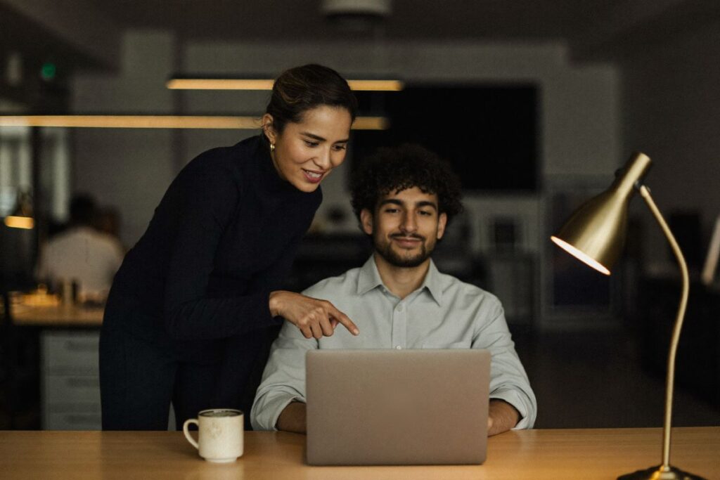 A manager points to a laptop as she welcomes a new hire at his desk on his first morning