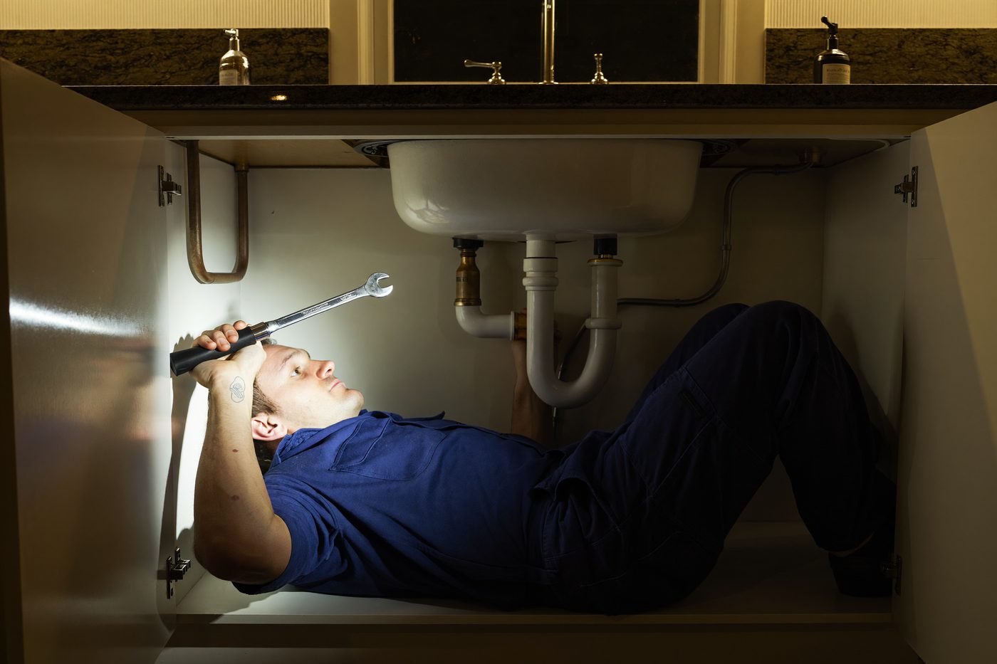 A plumber lies under a kitchen sink tightening a pipe fitting with a wrench, lit by a small flashlight