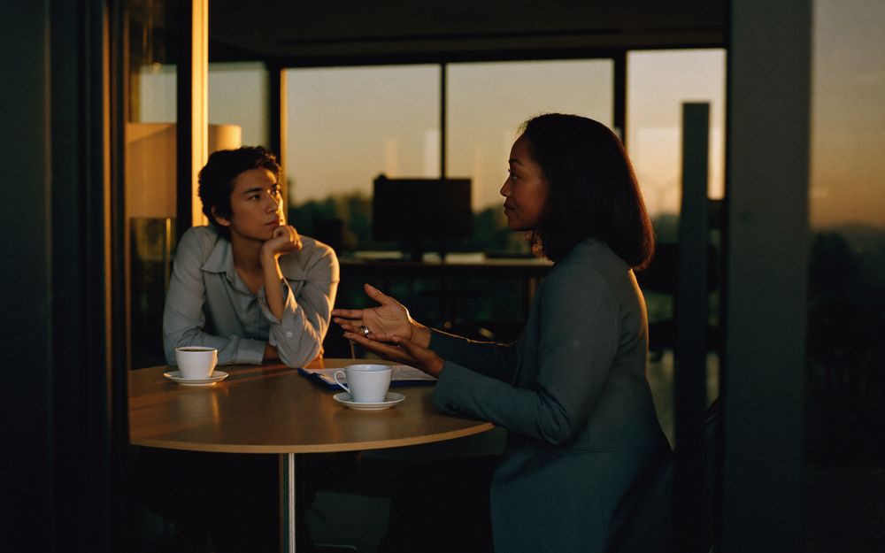 Two professionals in a candid interview at a small round table in a glass walled meeting room at sunset