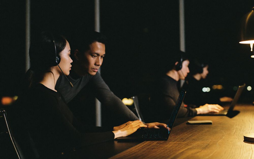 A team lead leans in beside a specialist wearing a headset to look at her laptop together, with two more specialists working along the desk